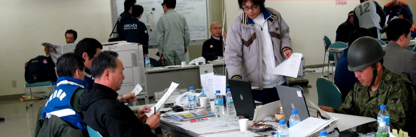 A disaster response coordination room — officials in emergency vests and a soldier working over laptops and paper reports.